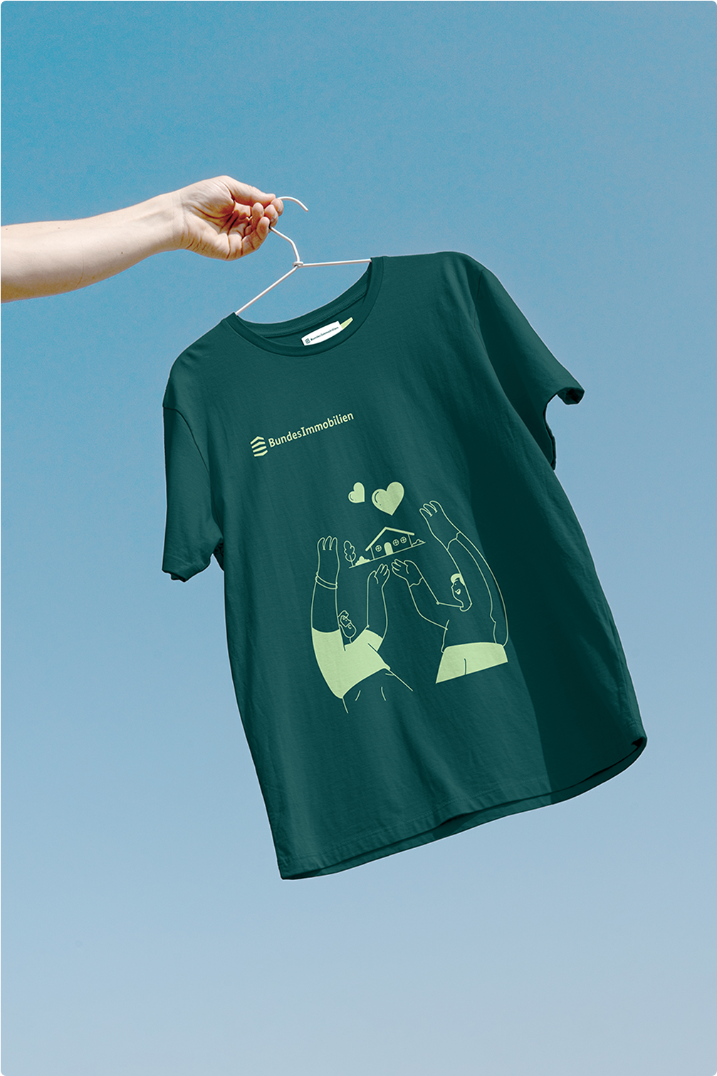 Eine Hand hält ein dunkelgrünes T-Shirt mit hellgrüner Illustration und Logo auf einem Kleiderbügel, vor blauem Himmel.