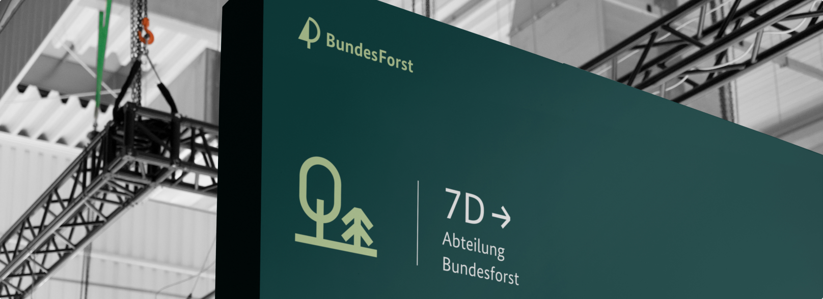 Ein Schild mit "BundesForst"-Logo und einem Icon. Darauf steht "7D → Abteilung Bundesforst". Im Hintergrund sind Teile einer Hallendecke zu sehen.