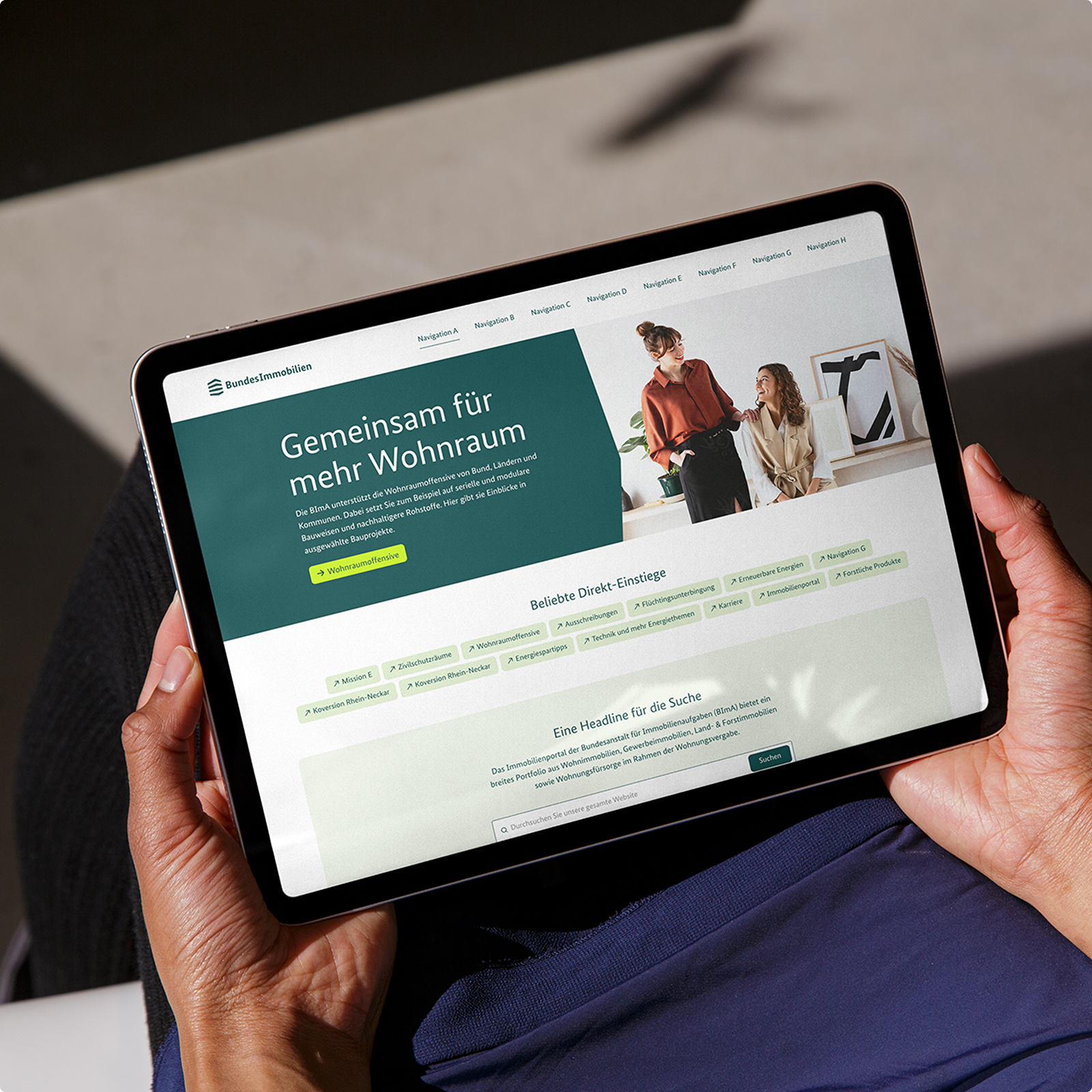 Eine Person hält ein Tablet, das die Website der Bundesimmobilien mit einem Titel, einem Bild und mehreren Navigationsoptionen zeigt.